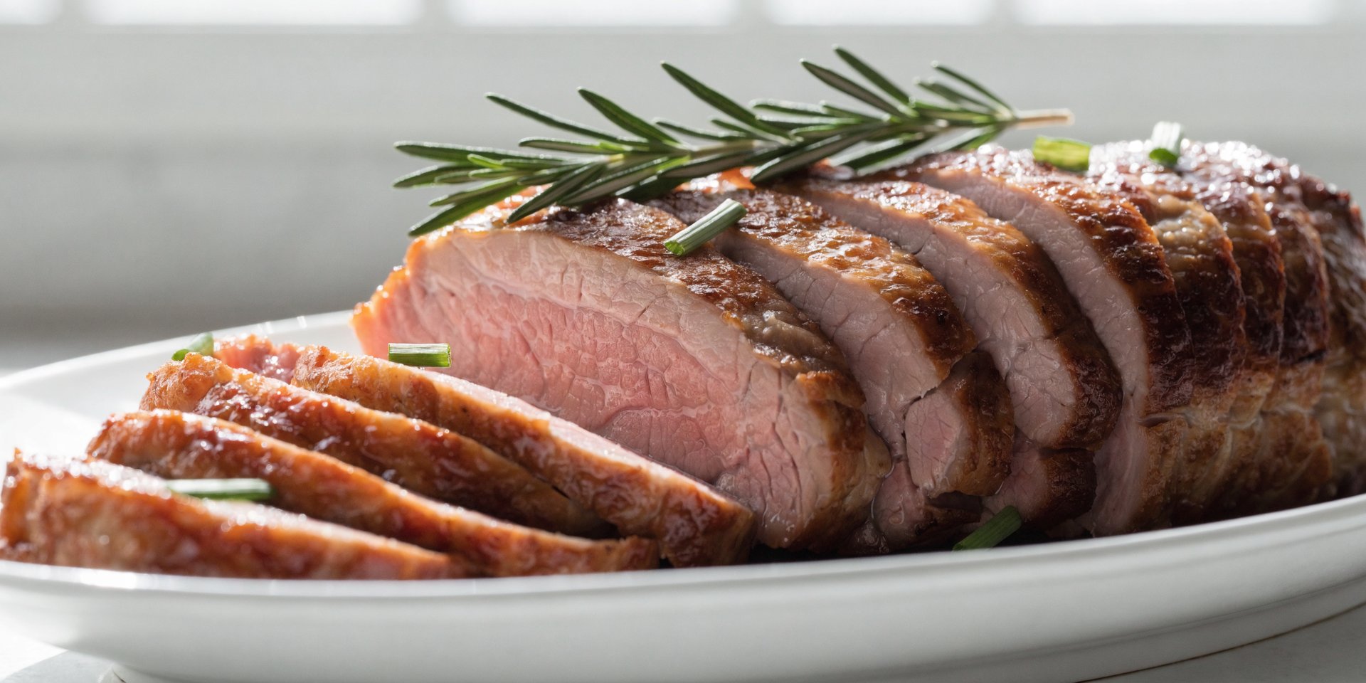 Slices of golden-brown pork tenderloin are fanned out on a white platter, garnished with rosemary and chives.