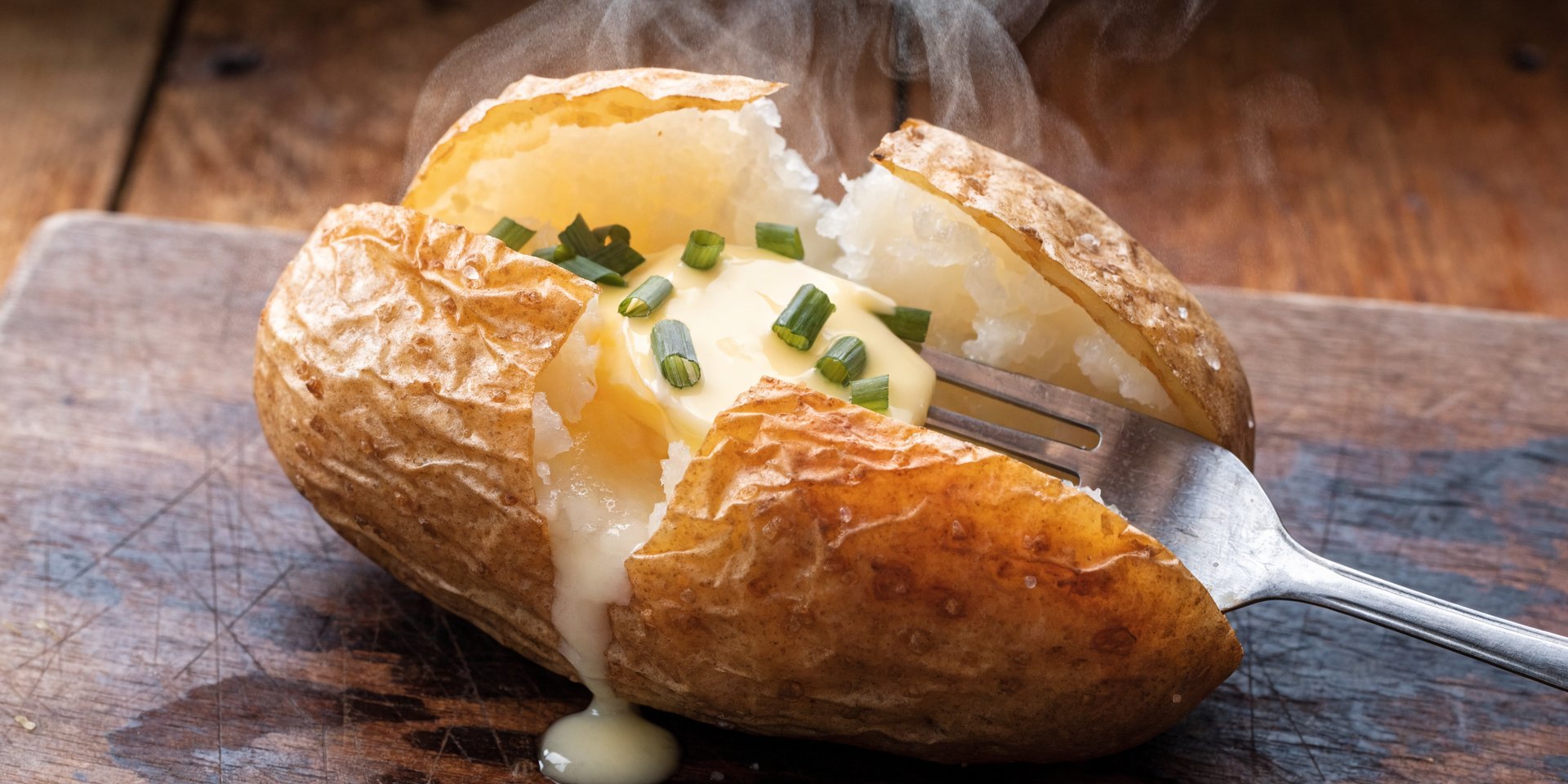 A perfectly baked russet potato is split open on a wooden board with melting butter and chives inside.