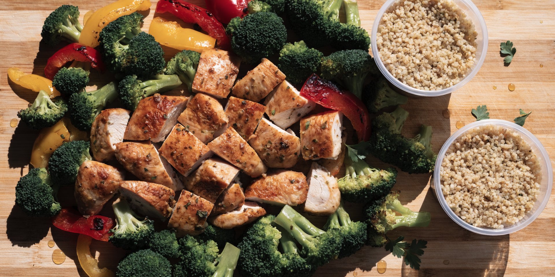 An overhead shot shows roasted chicken, colorful bell peppers, and broccoli florets arranged on a wooden cutting board with containers of grains.