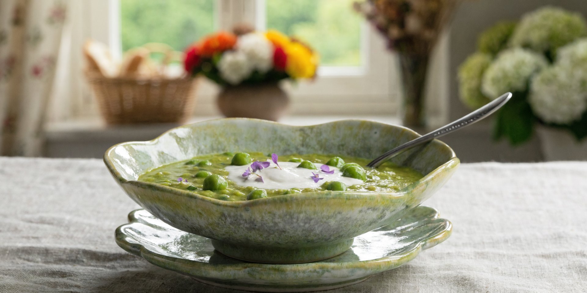 A bowl of bright green spring soup with a cream swirl and edible flower garnish sits on a light-colored surface.
