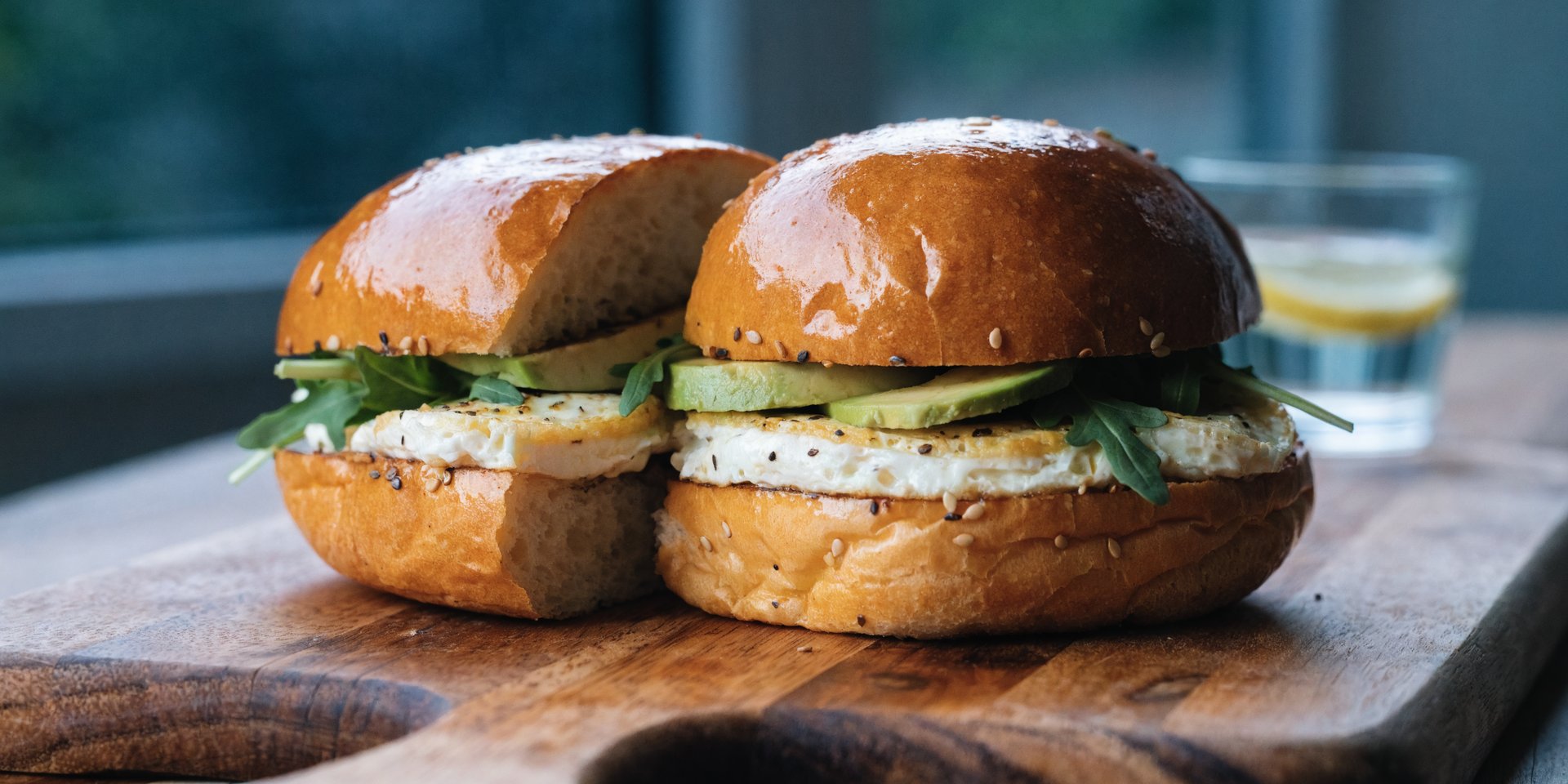 A healthy egg sandwich is cut in half, stacked with egg, arugula, and avocado, on a kitchen counter.