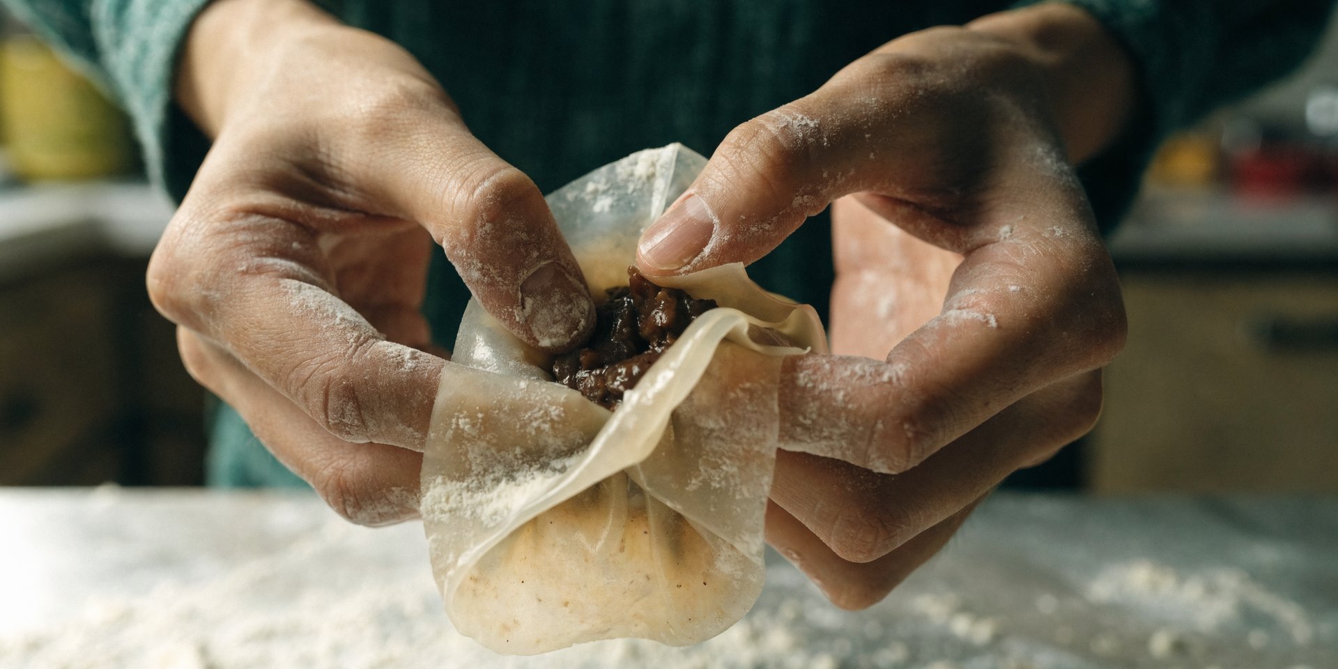 A pair of hands folds a round, thin sheet of dough around a dumpling filling on a lightly floured wooden surface.