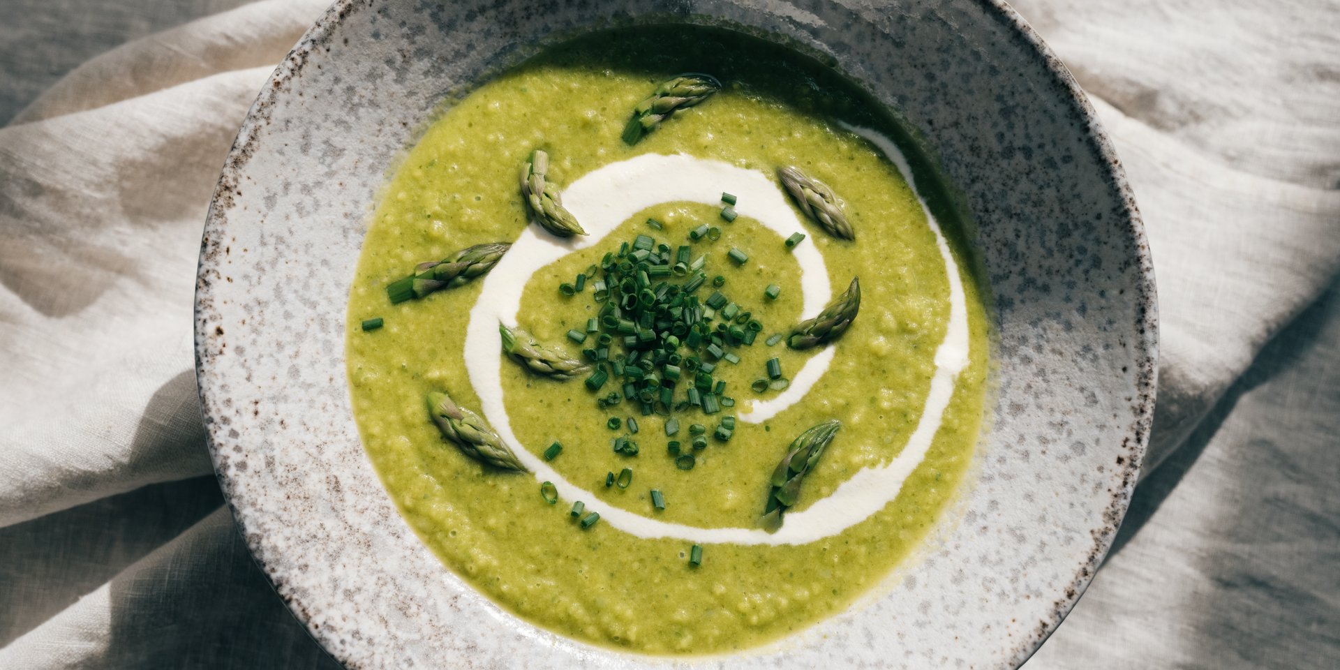 A vibrant green pureed soup in a rustic ceramic bowl is garnished with cream, chives, and asparagus tips on a light linen tablecloth.