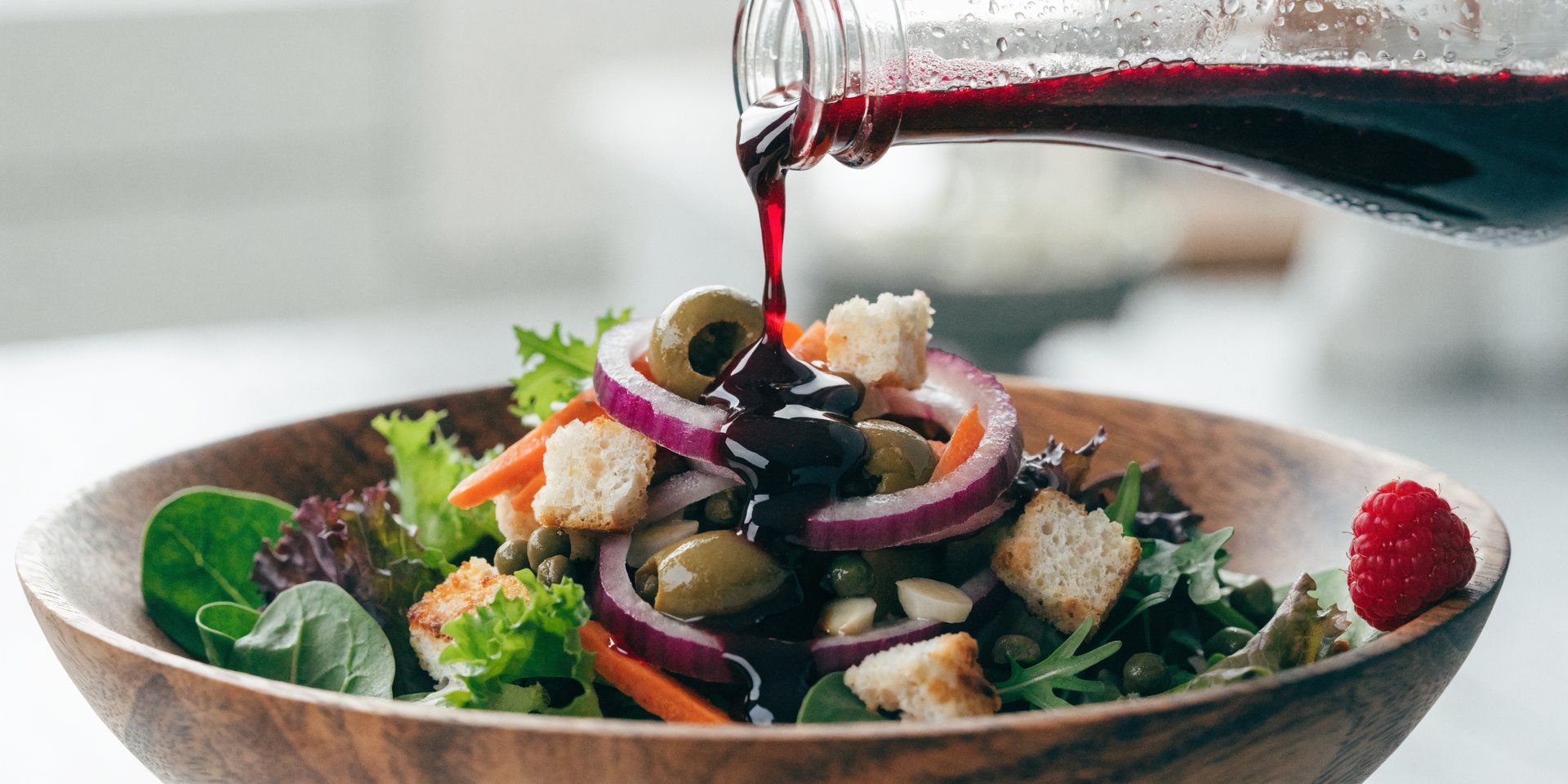 A close-up photograph shows a healthy green salad in a wooden bowl with a thick, glossy red salad dressing being poured over it.