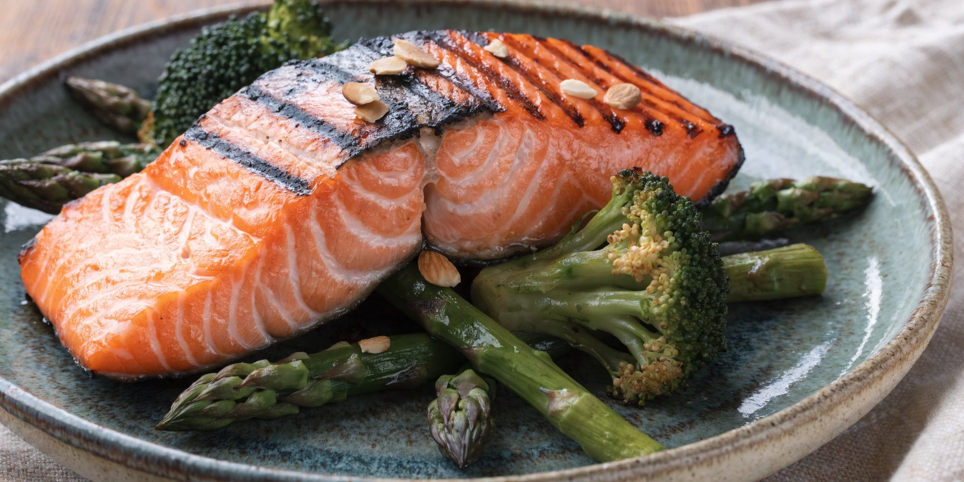 A grilled salmon fillet is plated with roasted broccoli, green asparagus spears, and toasted almonds on a ceramic dish.