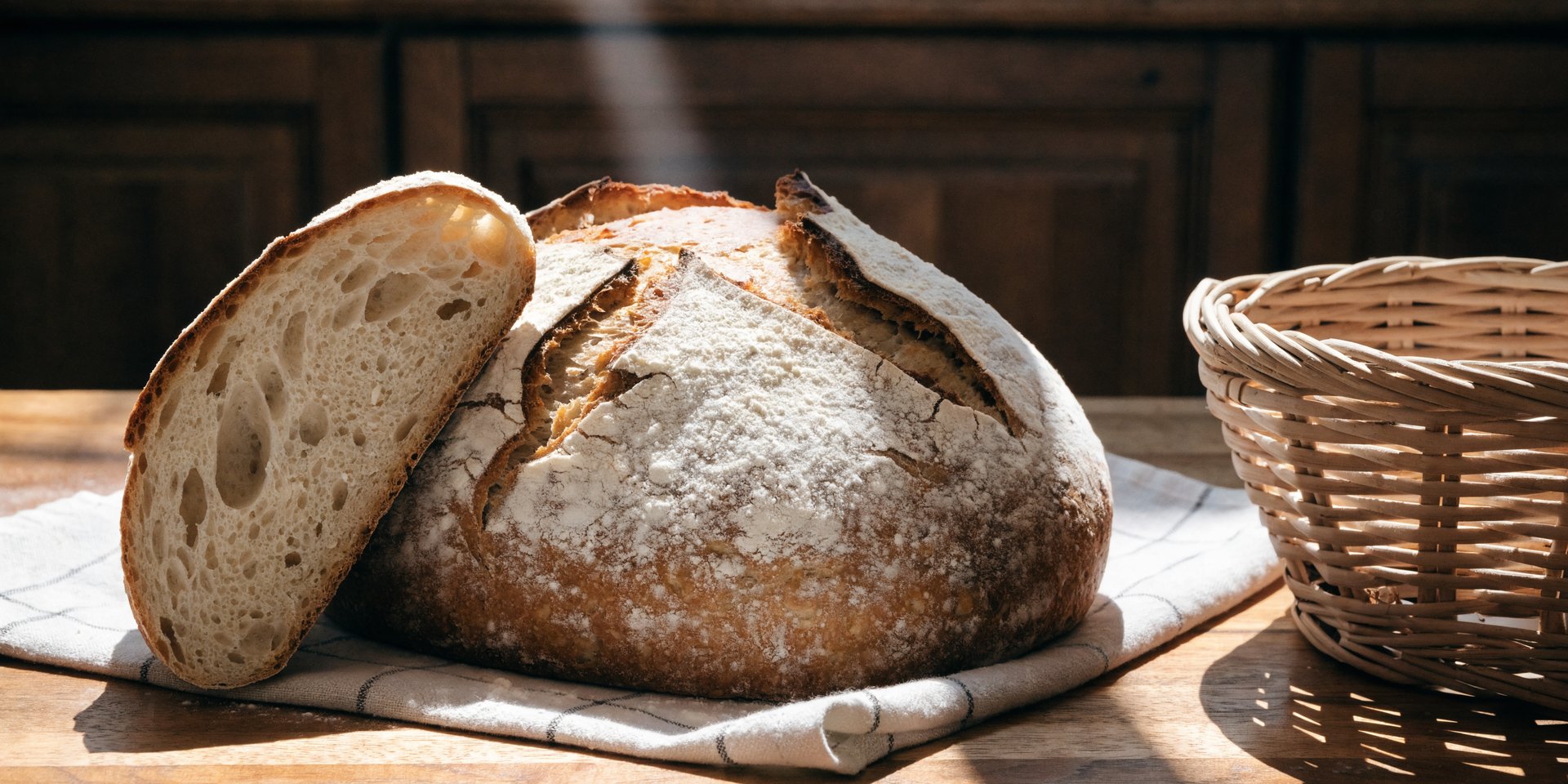 A golden-brown, round sourdough loaf with one slice cut sits on a light linen on a wooden surface.