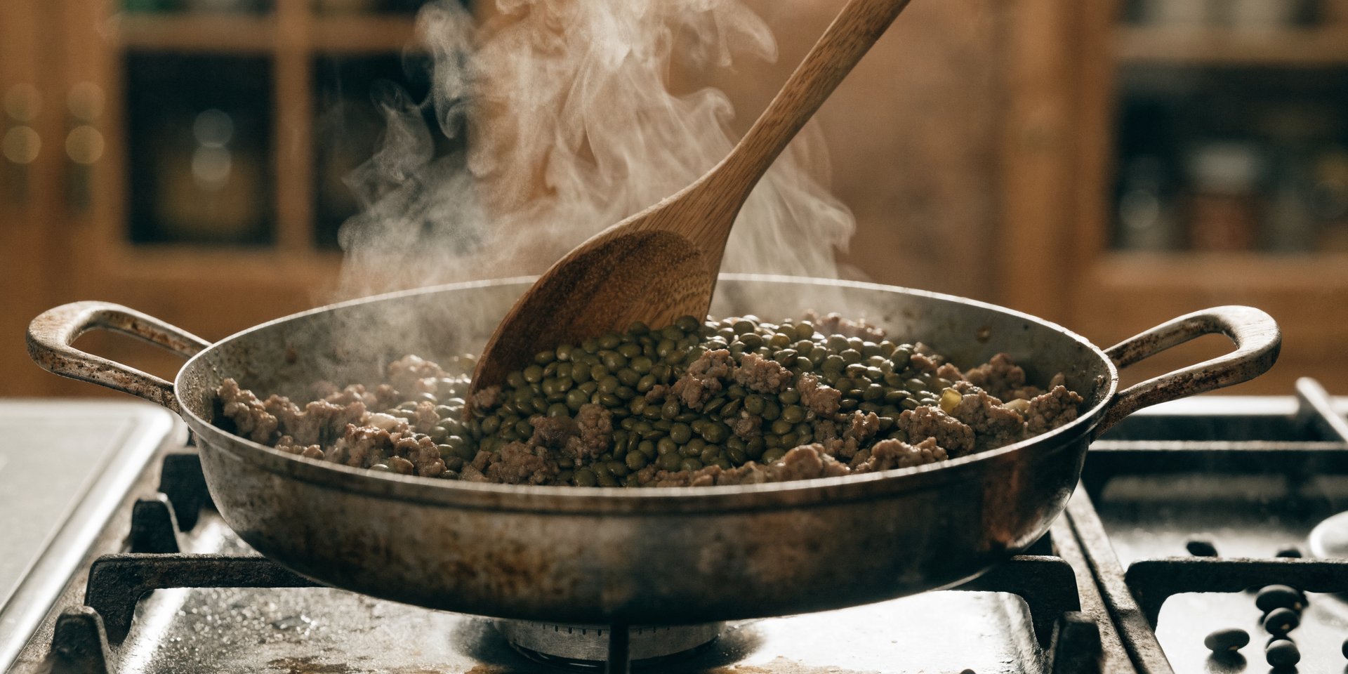 A close-up of a wooden spoon stirring ground meat and lentils in a cast iron skillet.