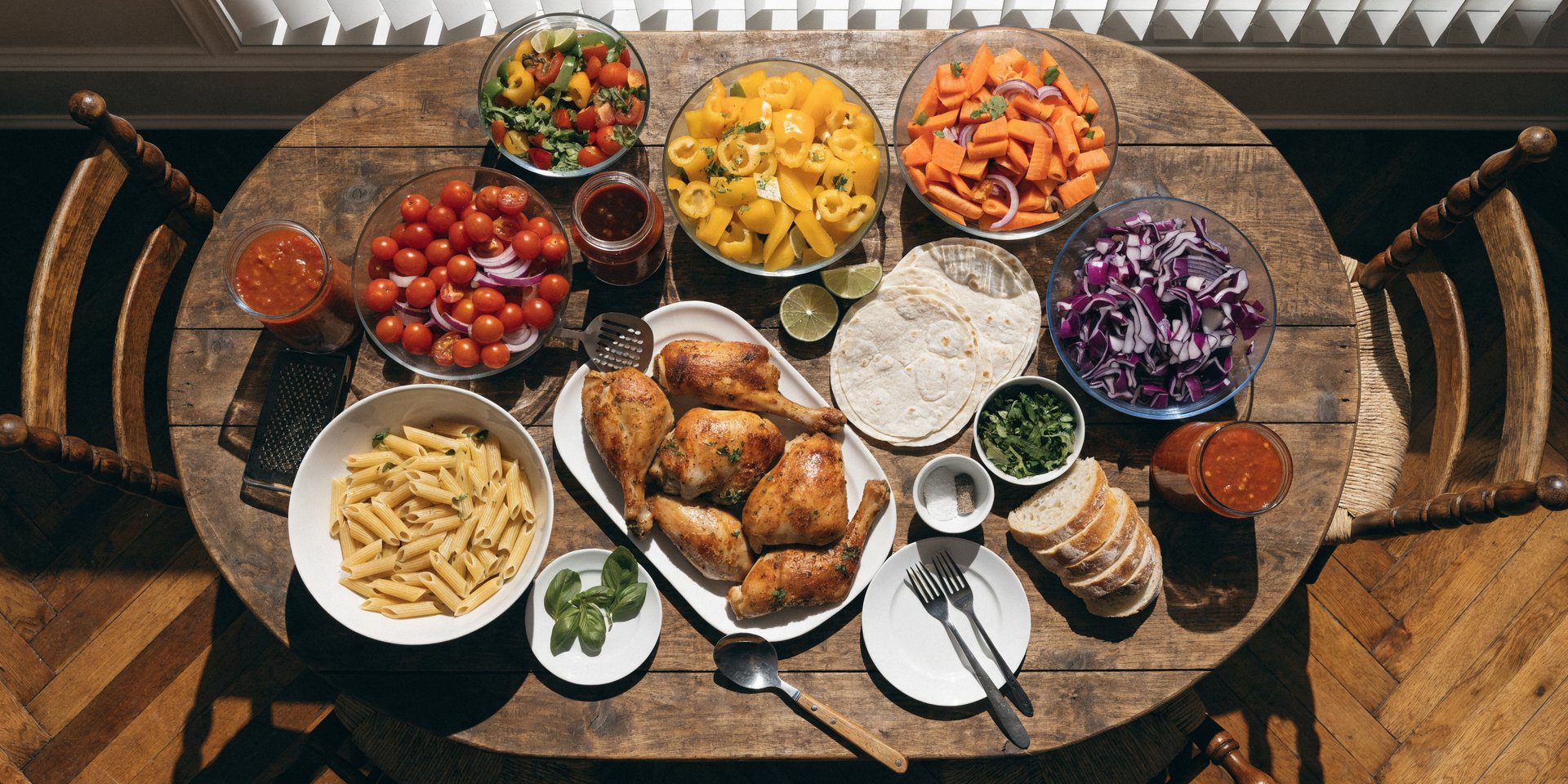 An overhead view of a large rustic wooden table laden with various prepared meal components like chopped vegetables, roasted chicken, pasta, and sauces.