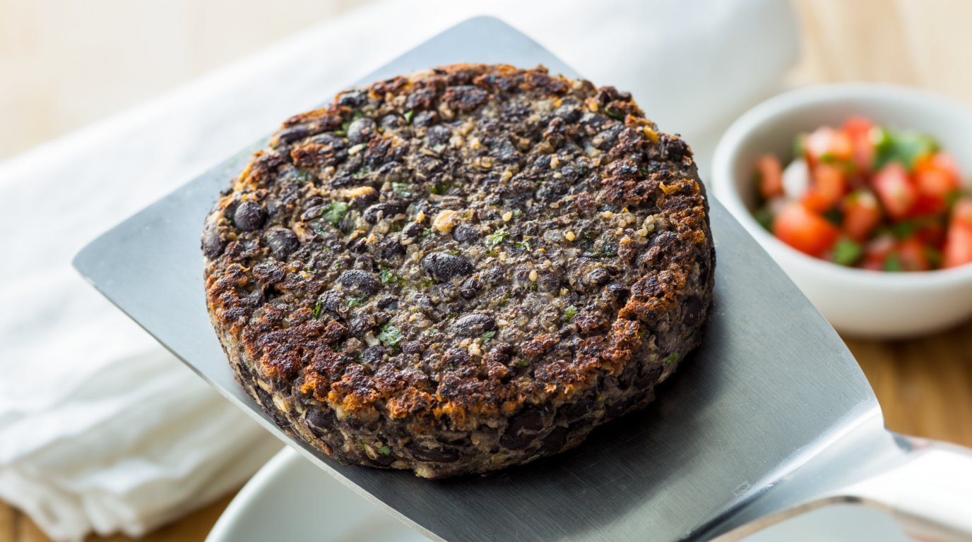A whole black bean burger patty held on a metal spatula with visible bean and grain texture on its intact surface.