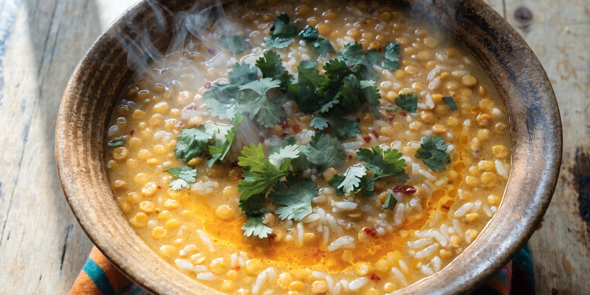 An overhead shot of a bowl of golden dal and rice, garnished with fresh cilantro and red chili flakes, on a wooden surface.