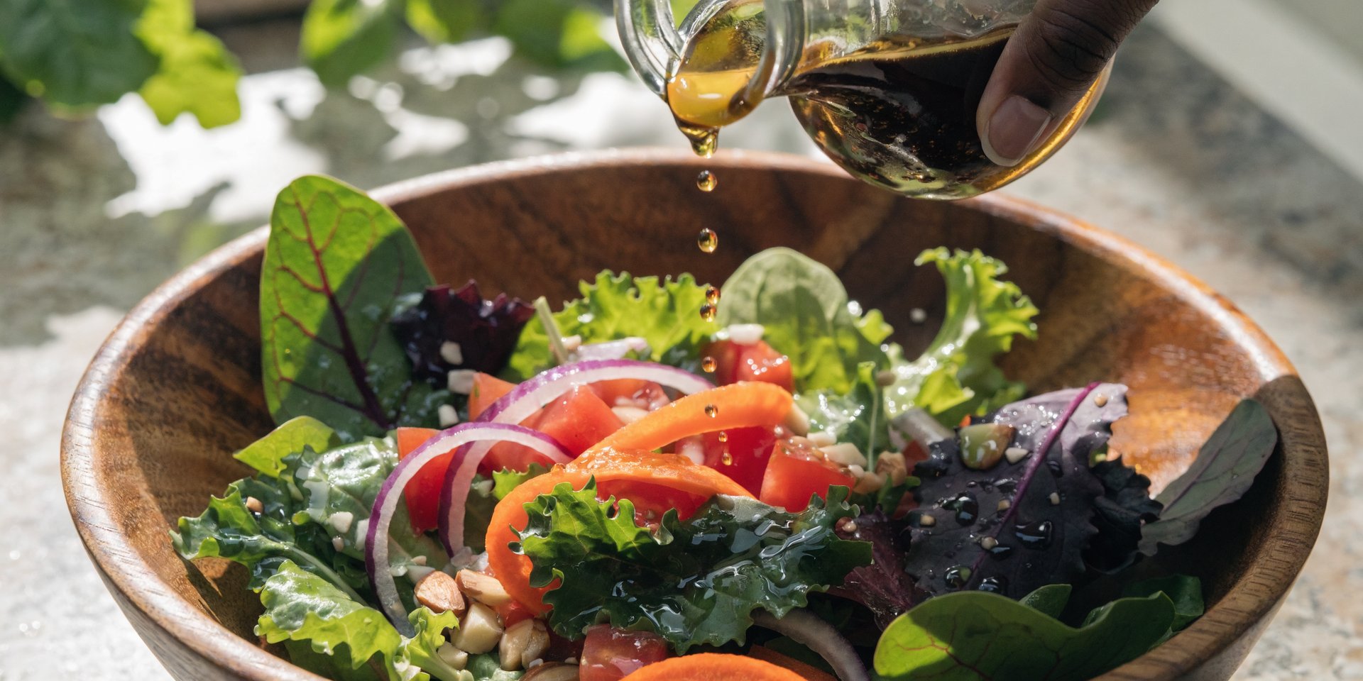 A hand drizzles homemade vinaigrette over a fresh green salad in a wooden bowl.