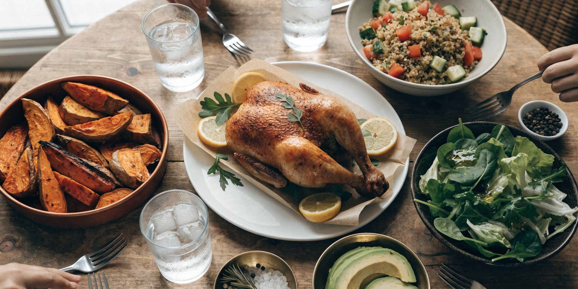 An overhead shot of a balanced family dinner featuring roasted chicken, roasted sweet potatoes, a green salad, and avocado slices on a rustic wooden table.